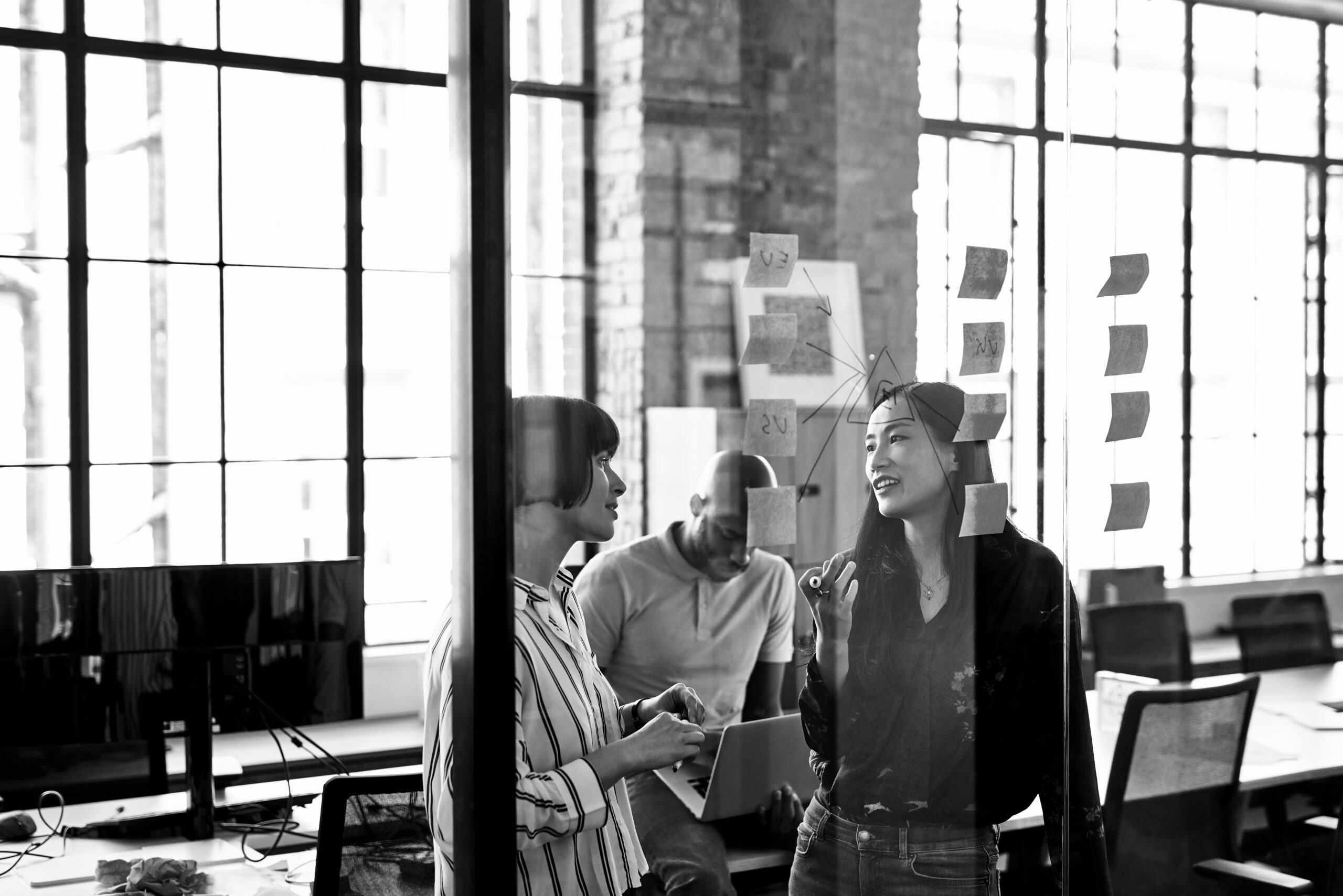 Three people collaborate in a modern office. Two women stand discussing by a glass wall with sticky notes, while a man works on a laptop in the background.