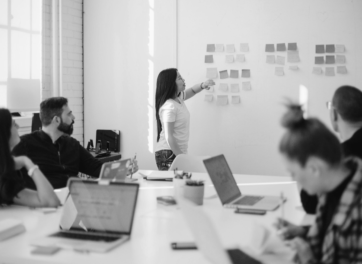 A woman points to a whiteboard covered in sticky notes while addressing four colleagues seated around a table in a modern office.