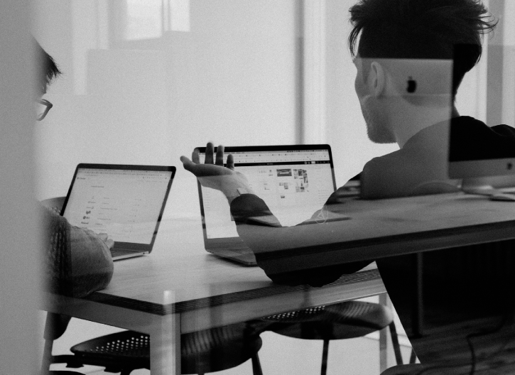 Two individuals sit at a table, each using a laptop. One person is gesturing towards their screen, while the other looks on. They appear to be in a discussion. The scene is monochromatic.