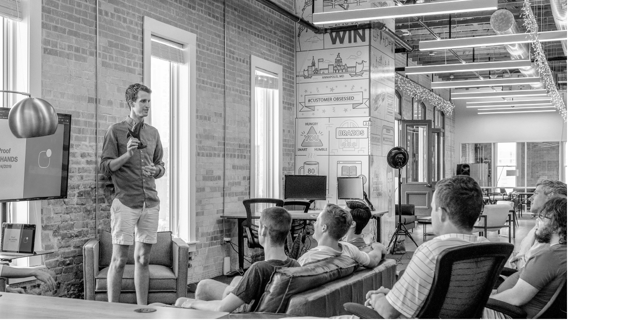 A person stands in front of a group of seated individuals, giving a presentation in an office space with brick walls and motivational posters.